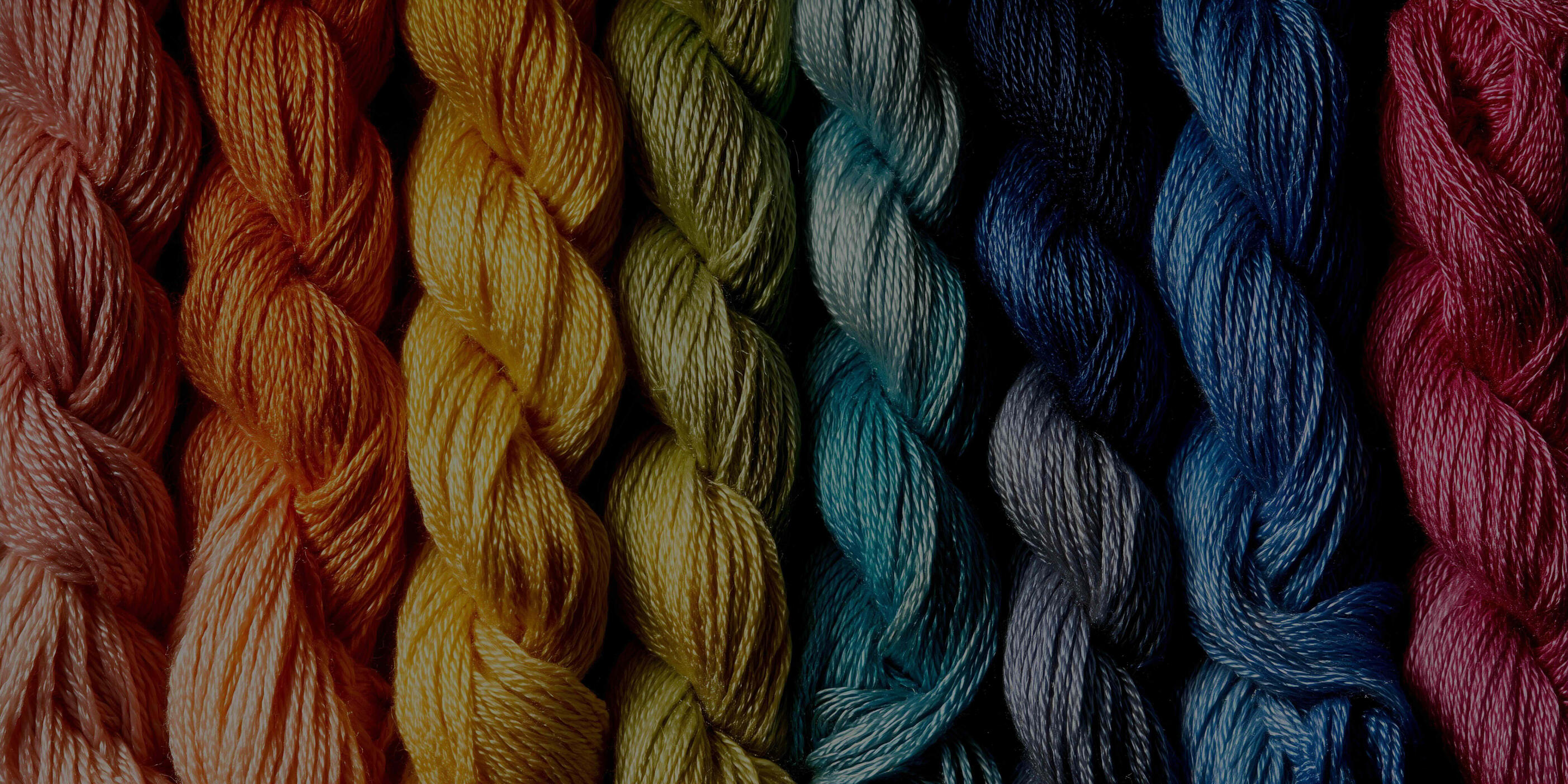Colorful Needlepoint Thread Skeins Close-up of colorful silk thread skeins arranged side by side in shades of coral, orange, yellow, olive green, teal, navy, blue, gray, and pink.